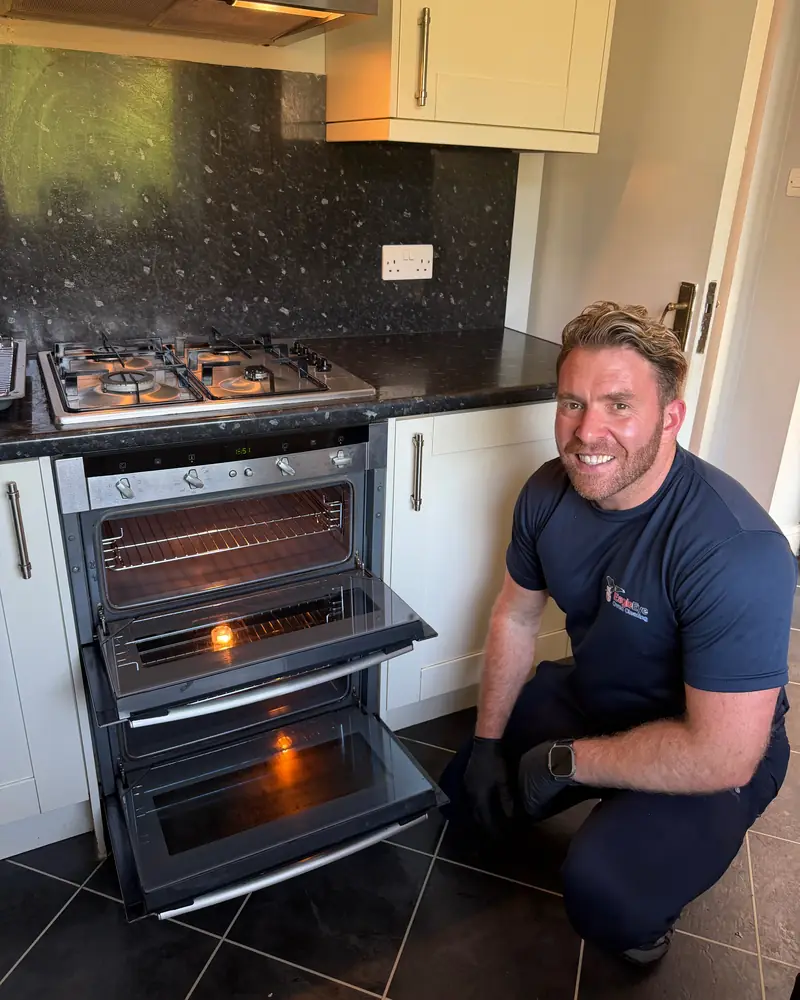 Cameron, founder of Eagle Eye Oven Cleaning, kneeling beside a clean double oven in a customer's kitchen, wearing an Eagle Eye t-shirt